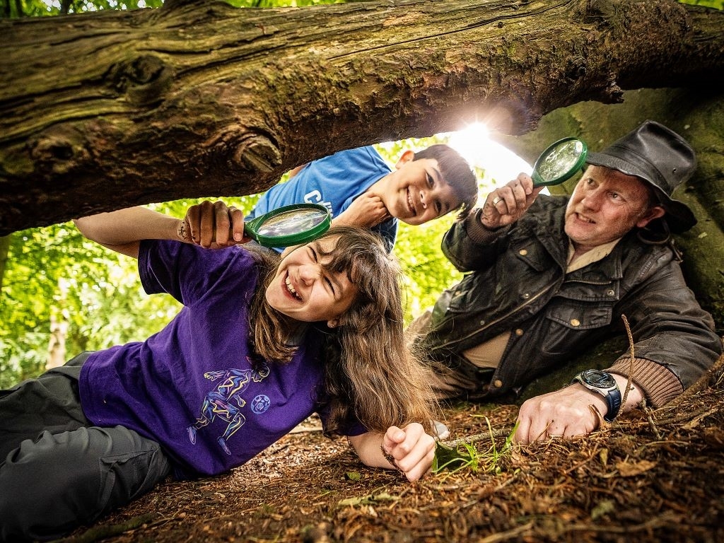 Two children looking under a tree branch with magnifying glasses, guided by a staff member.