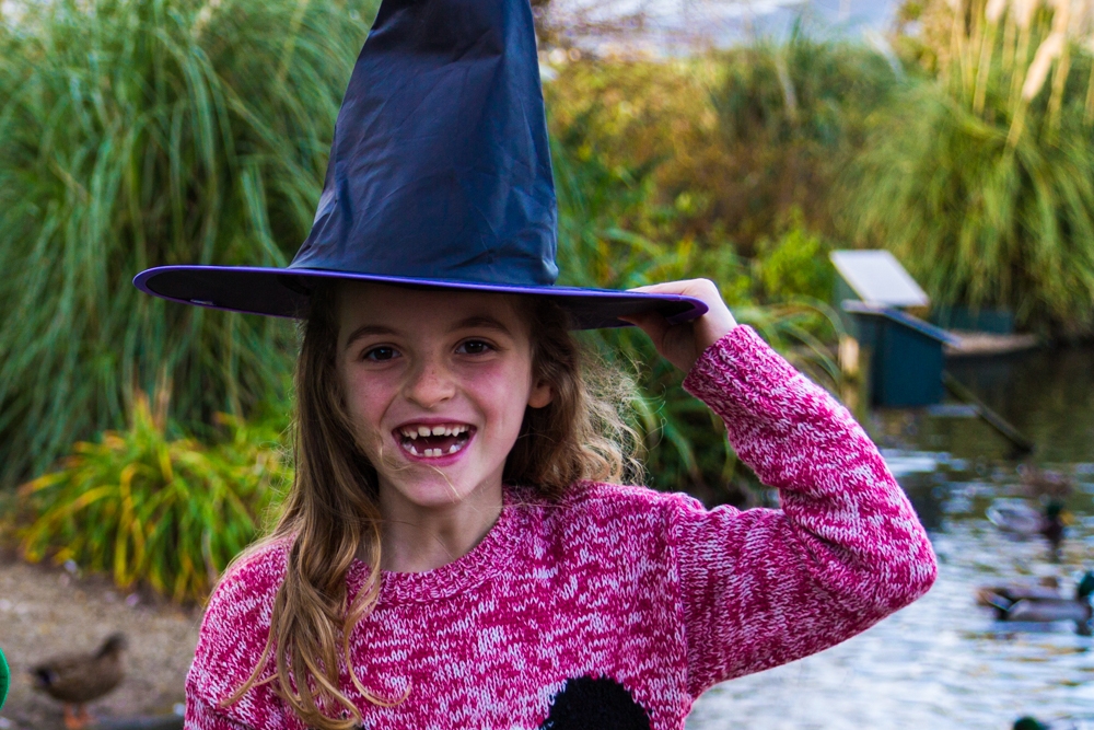 A girl smiling at the camera and holding onto a witches hat she is wearing.