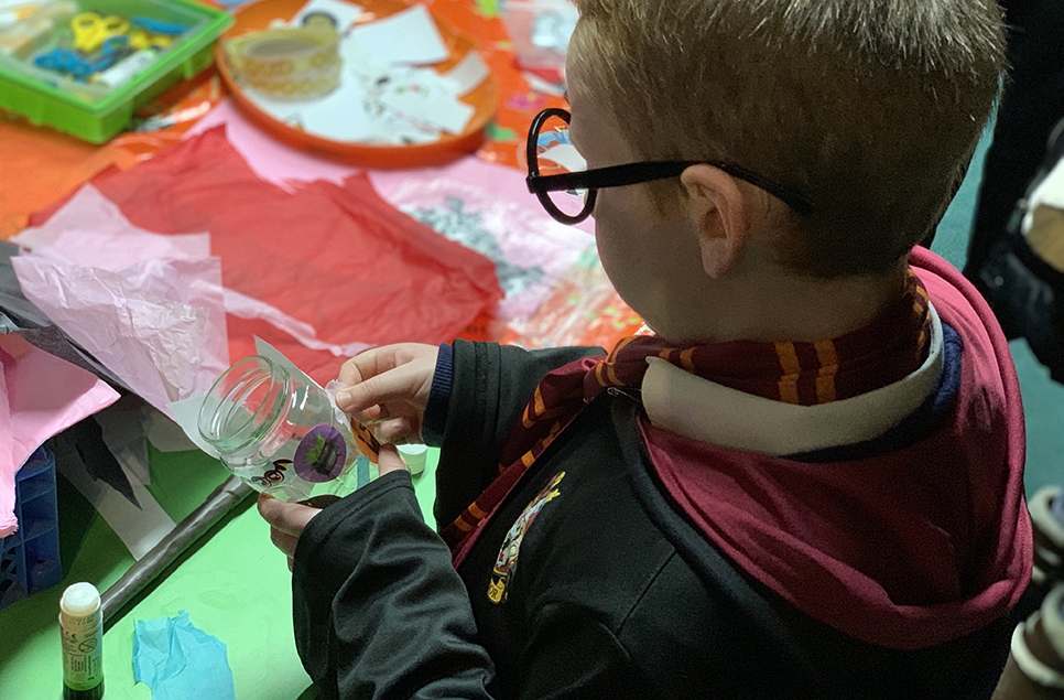 A little boy dressed up in Halloween Costume making a Halloween lantern from a jam jar.