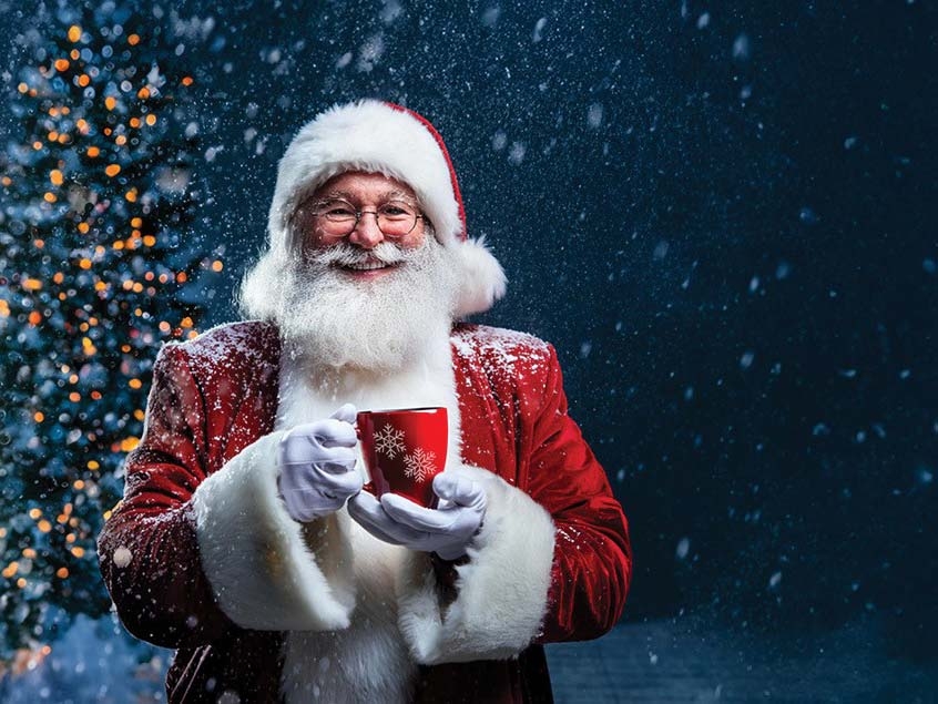 Santa smiles into the camera with a mug of warm drink in his hands.