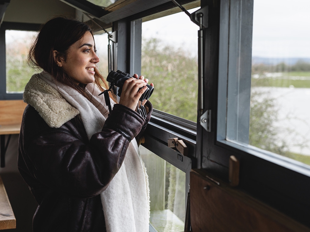 A visitor in a hide, smiling while looking out at the wetlands, with binoculars in their hands