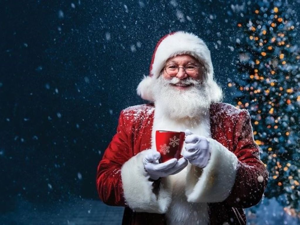 Santa Claus holding a festive mug and standing in front of a Christmas tree. Snow falls in the background.